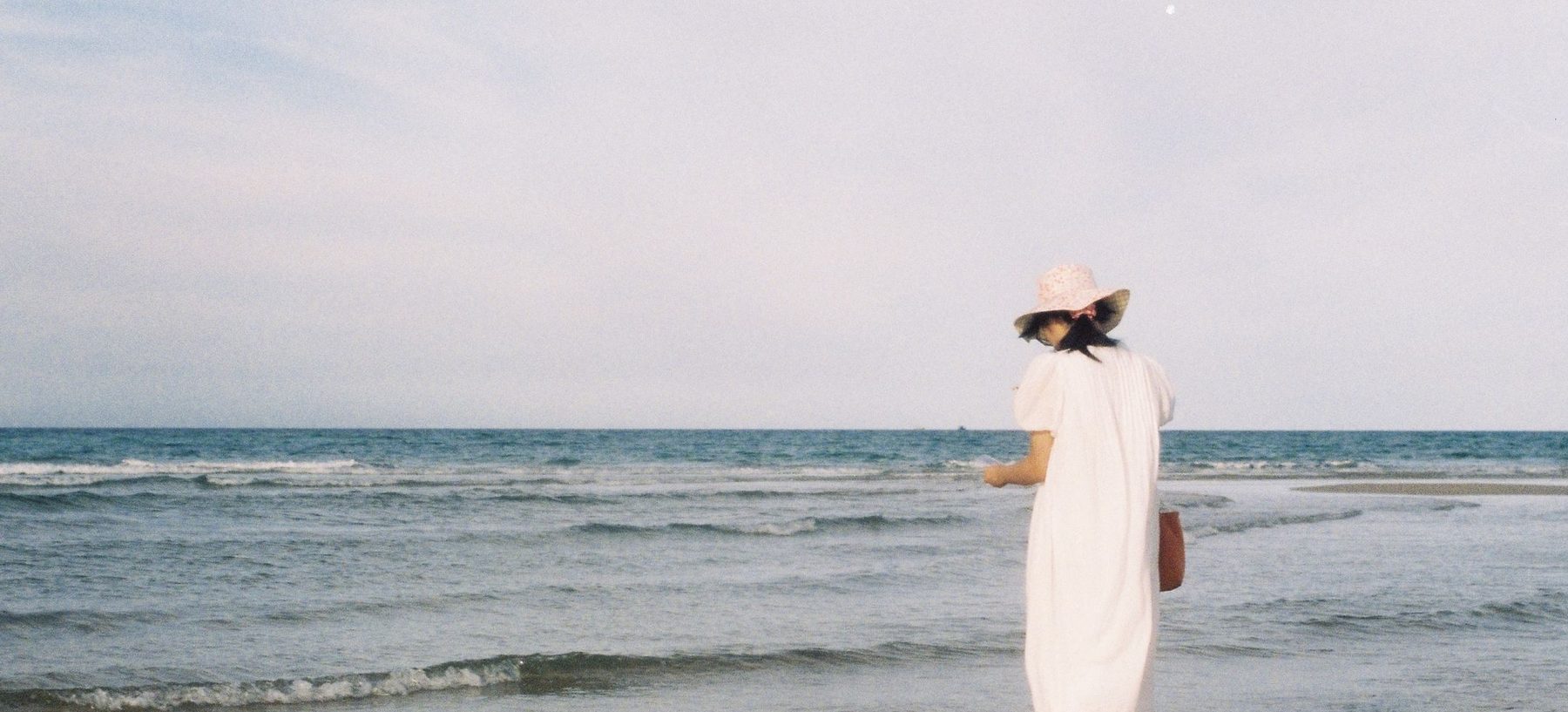 A person in a long white dress and a sun hat standing at the water's edge under a wide pale sky