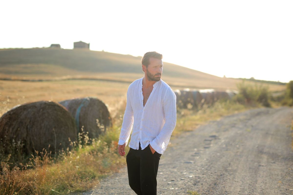 A man in a white linen shirt walking along a dirt road between round bales in evening light